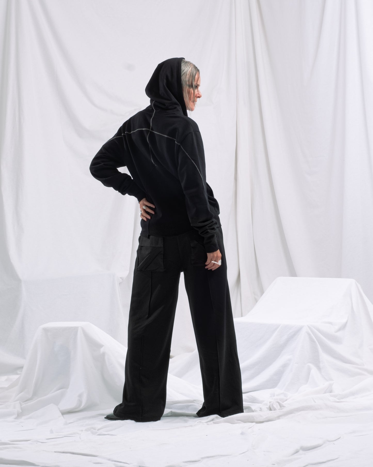 Person steht mit dem Rücken zur Kamera in einem weißen Studio und trägt eine schwarze Jogginghose mit weitem Bein und aufgesetzten Taschen. Die Hose ist kombiniert mit einem schwarzen Zip-Hoodie mit Kontrastnähten.