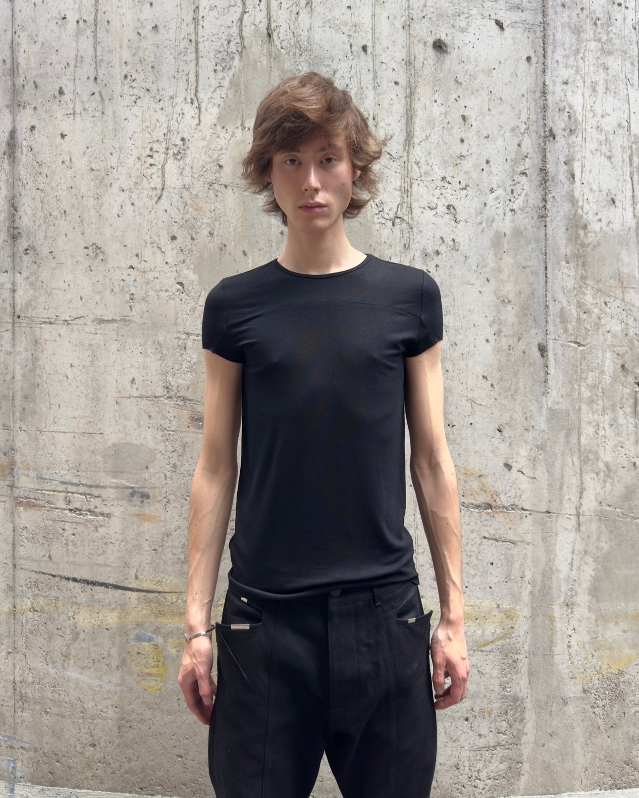 Person trägt das enganliegende schwarze Kurzarm-T-Shirt in Frontalansicht. Das Shirt sitzt körperbetont und zeigt die horizontale Teilungsnaht auf Brusthöhe. Die Person trägt eine schwarze Hose.