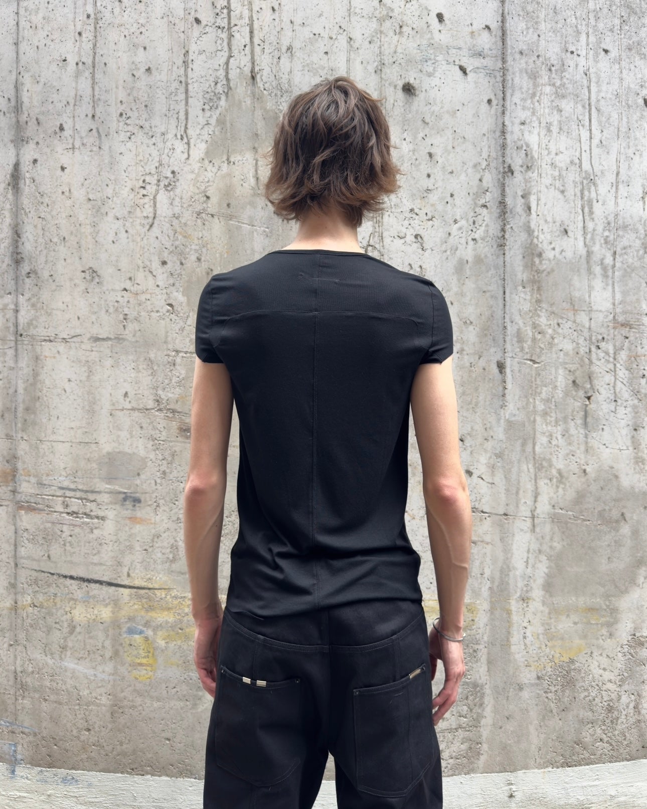 Rückansicht der Person im eng anliegenden schwarzen T-Shirt. Die vertikale Rücken-Naht ist gut sichtbar, ebenso wie die taillierte Passform. Die schwarze Hose sitzt locker an der Hüfte.